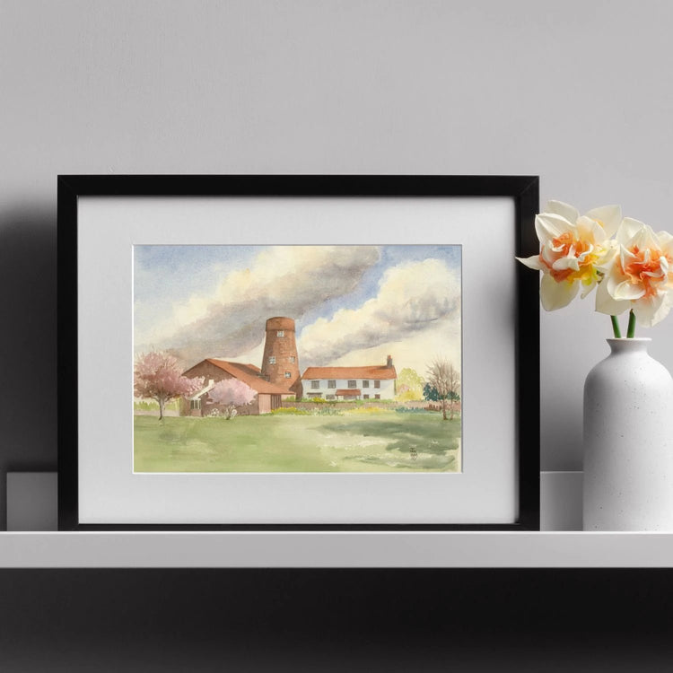 Framed and mounted mill print on a white shelf next to a small vase of white and orange flowers